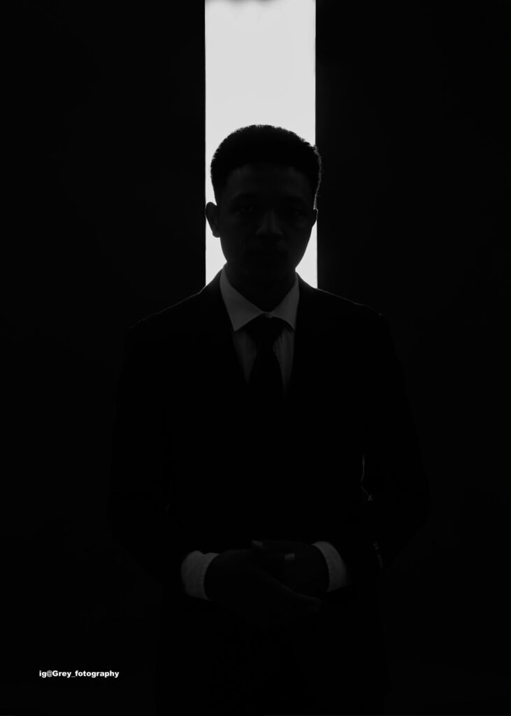 Low-key black and white portrait of Royking Niba in a dark suit and tie, backlit by a narrow vertical strip of light