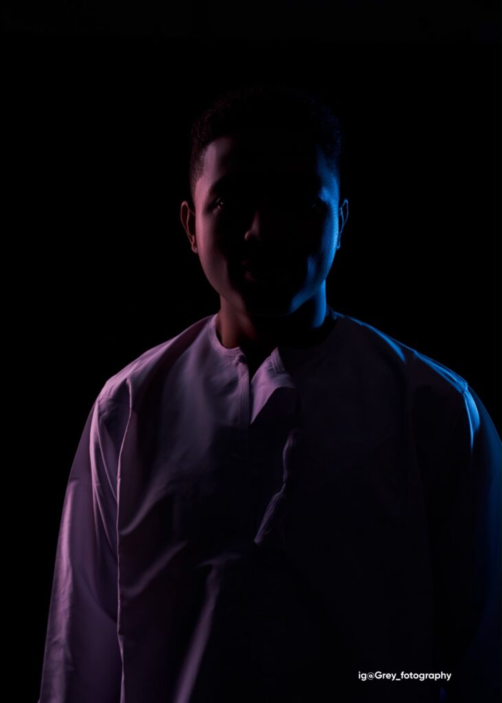 Portrait of Royking Niba in a white collarless shirt, lit from the side by blue and magenta light against a black background