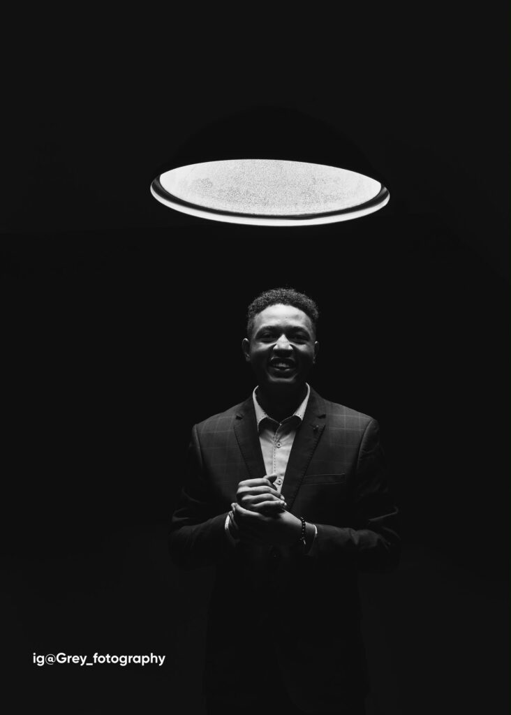 Black and white portrait of Royking Niba in a checked suit, smiling with hands clasped, beneath a circular ring light