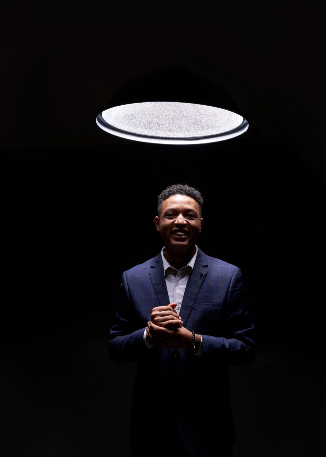 Royking Niba in a navy checked suit, hands clasped, standing beneath a circular ring light against a black background